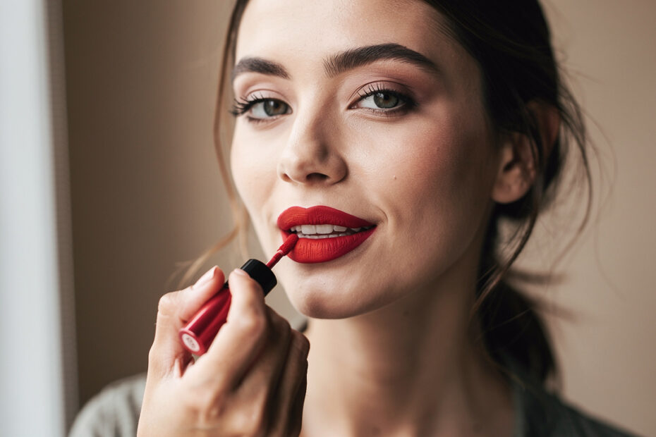 Portrait beauté élégant d’une jeune femme appliquant un rouge à lèvres liquide mat rouge à la lumière naturelle, sur un fond beige sobre.